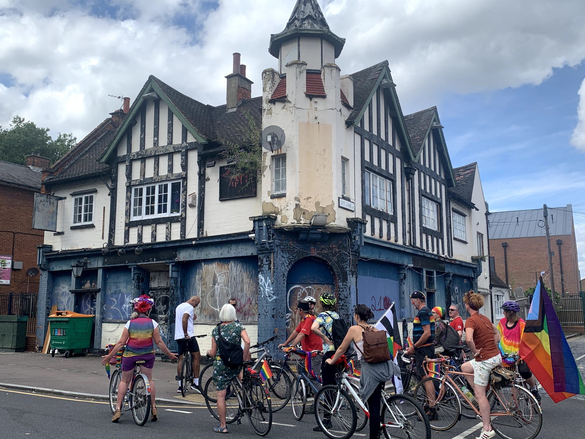A group of cyclists outside the semi derelict pub The Angel. Some of the riders wear or carry Pride flags