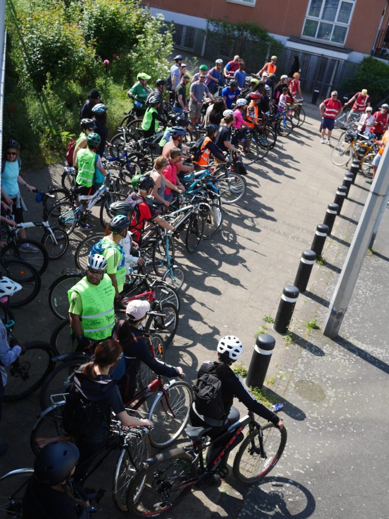 Lots of people with bicycles standing on a paved area (separated from the road by bollards) waiting to set off.